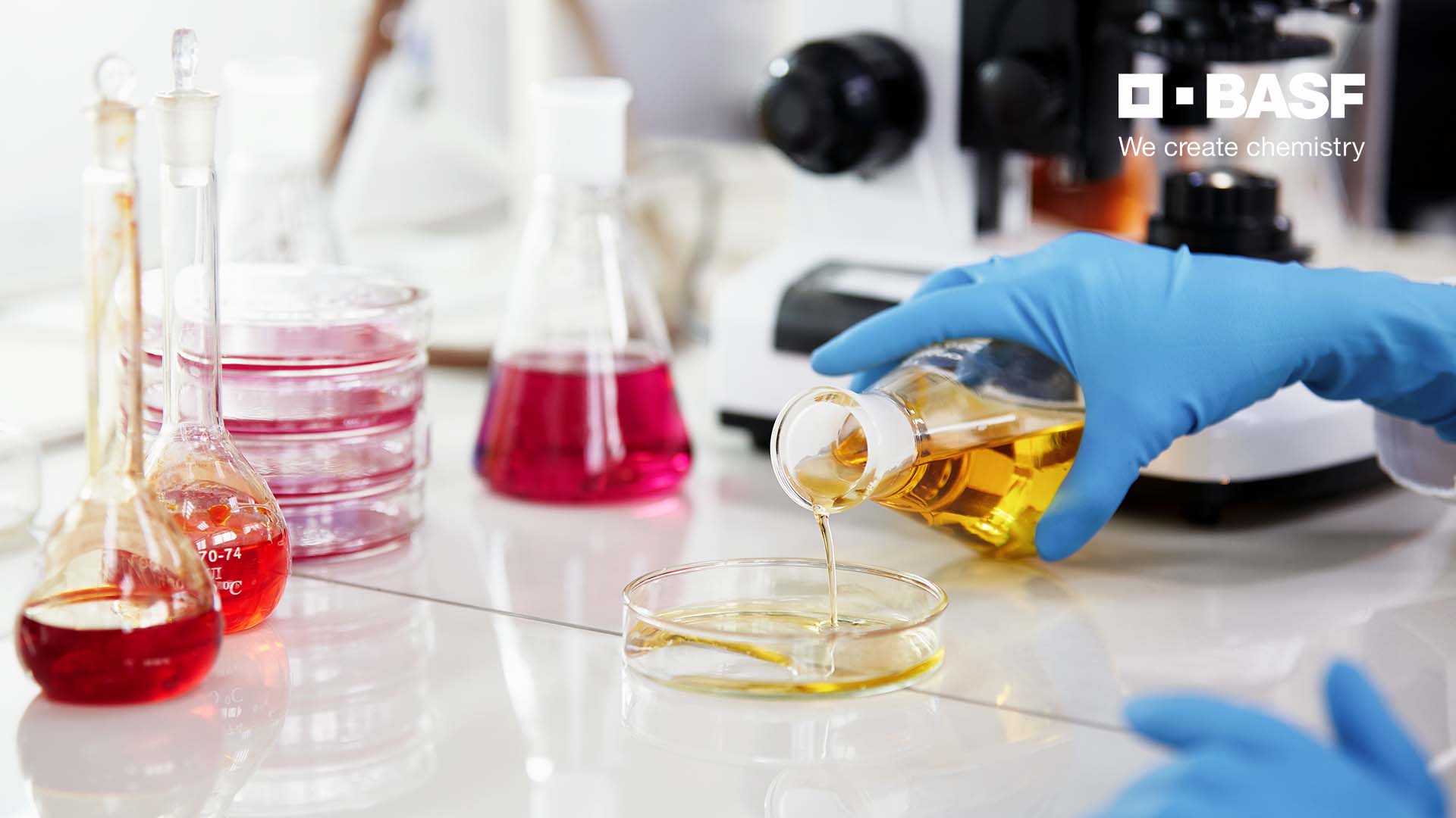Laborhände mit blauen Handschuhen gießen eine gelbe Flüssigkeit in eine Petrischale, BASF-Logo „We create chemistry"