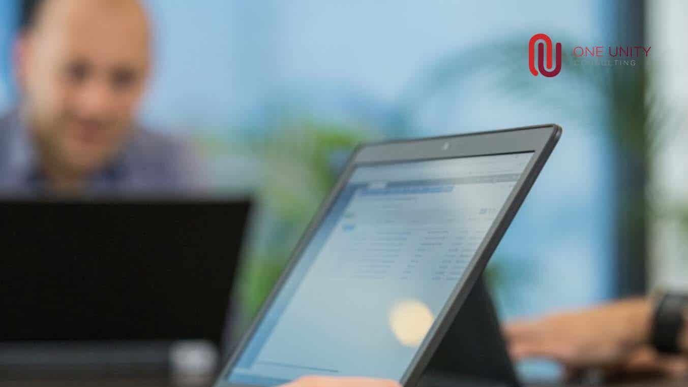 Person arbeitet an einem Tablet, im Hintergrund ein Laptop-Bildschirm, mit One-Unity-Consulting-Logo
