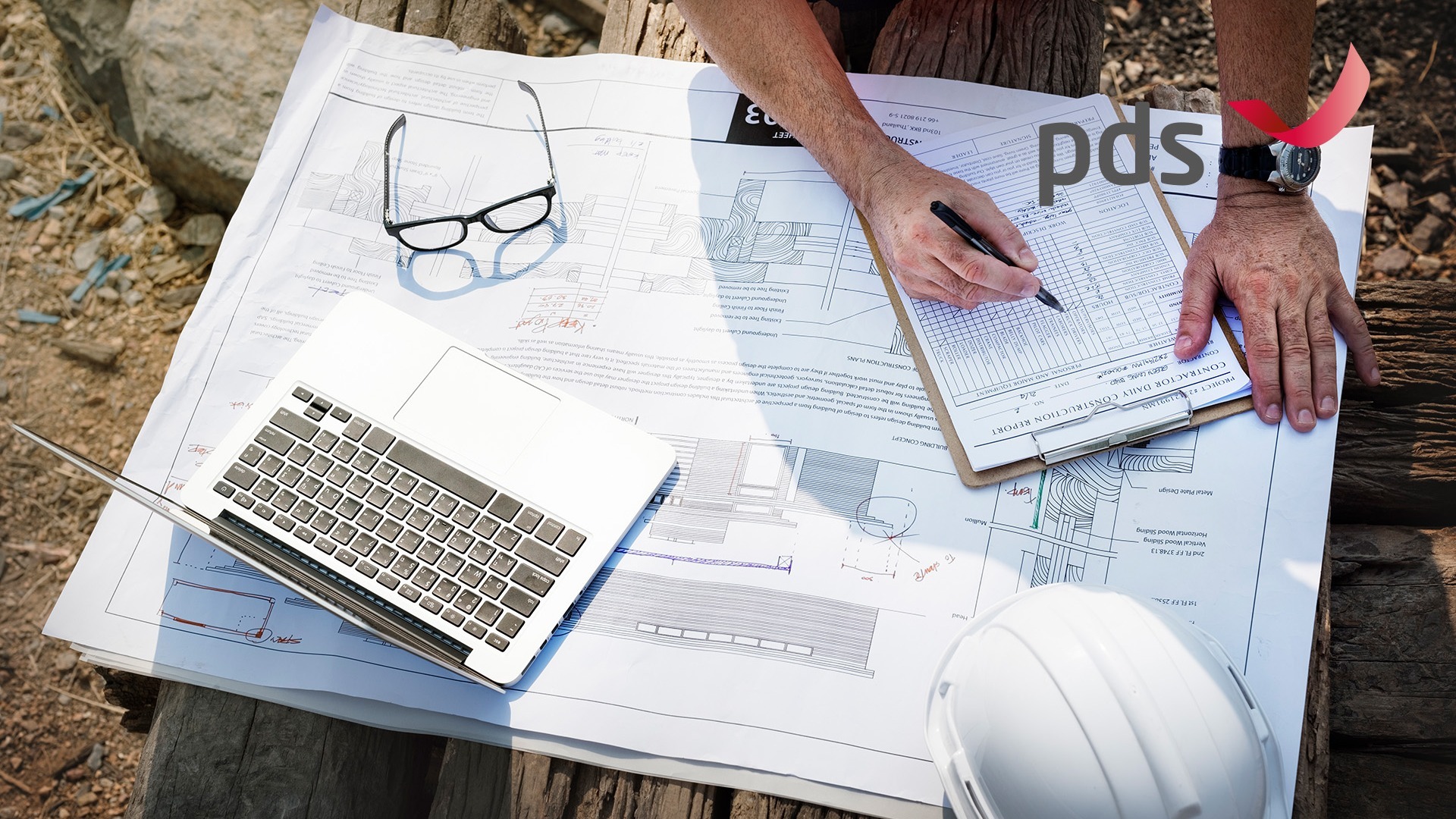 Blaupausen, Laptop, Brille und Bauhelm auf einem Holzstapel auf einer Baustelle, mit pds-Logo